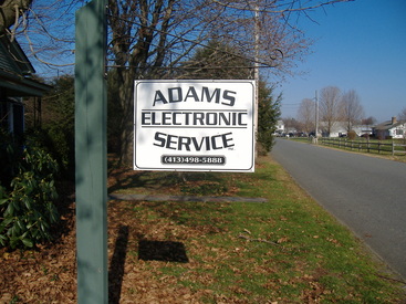 The Adams Electronic Service roadside sign, lettered with the phone number 413-498-5888, on a post beside a country road.