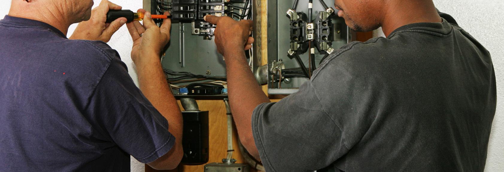Two electricians working side by side inside an open electrical panel and meter stack.