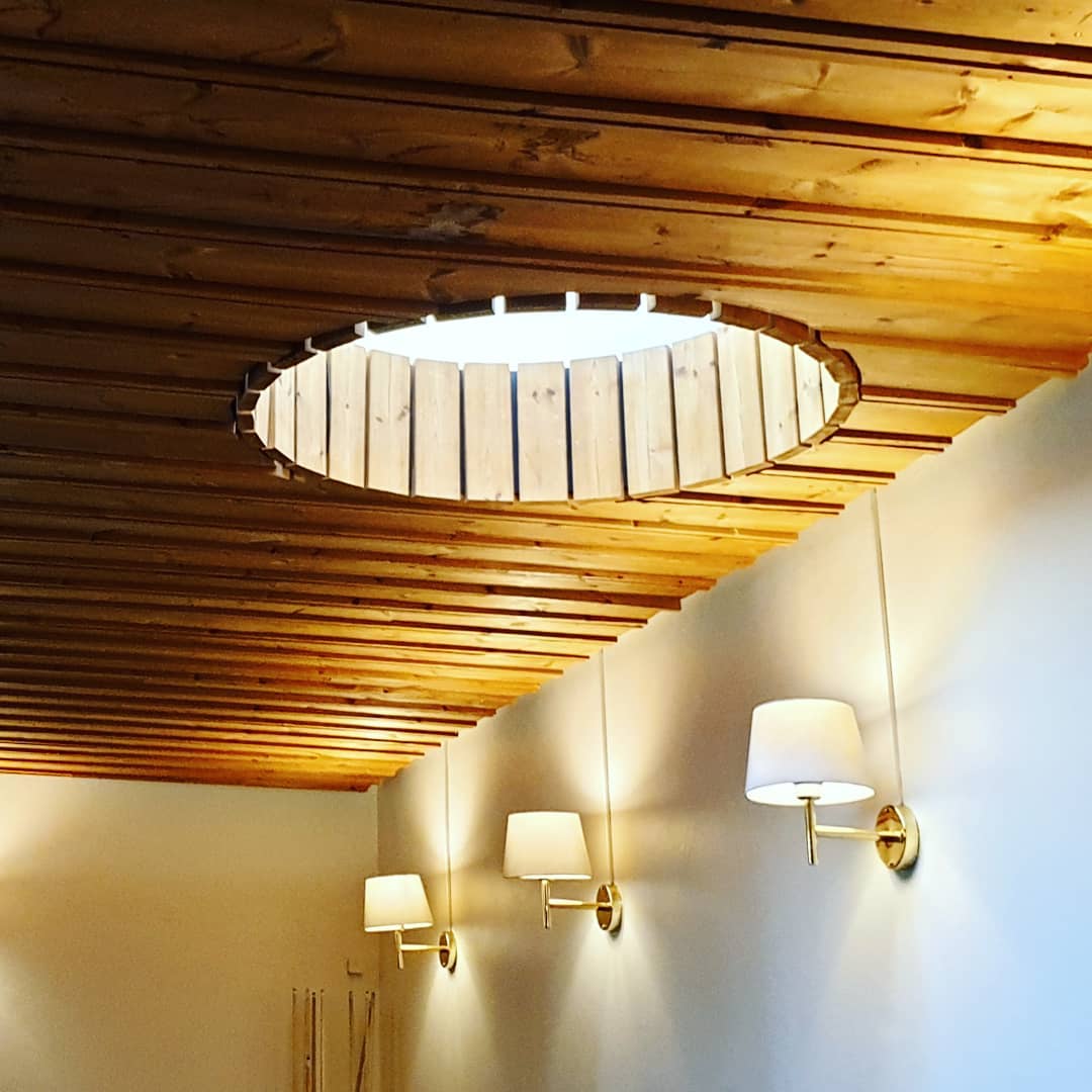 A warm-lit room with a slatted wood ceiling, a round light cove cut into it, and three brass wall sconces lit below.