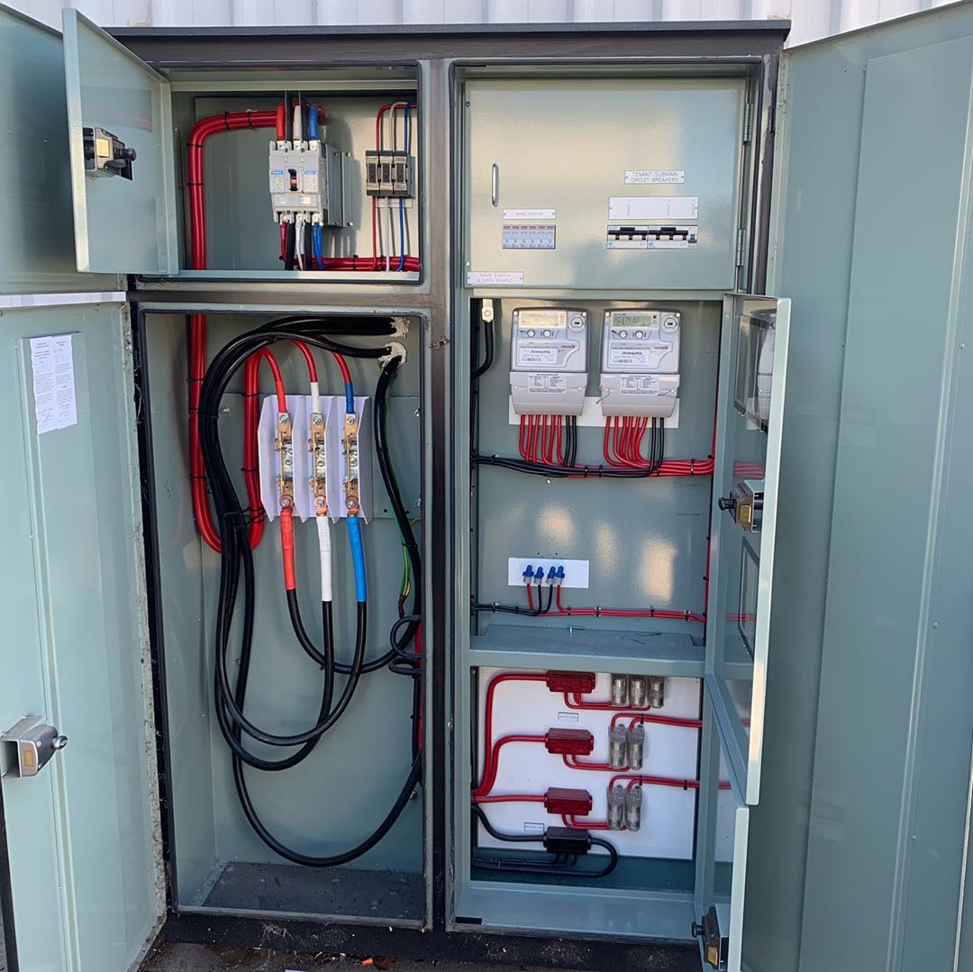 A green control cabinet standing open, with breakers, meters, fuses and red, blue and black conductors inside.