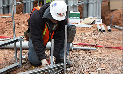 An electrician in a hard hat and high visibility vest assembling strut and conduit supports on a site