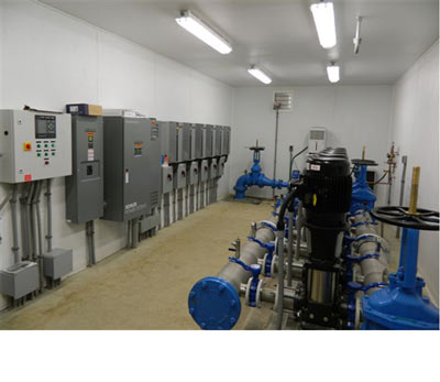 A row of control panels and drives along the wall of a pump room with pumps and valves on the floor