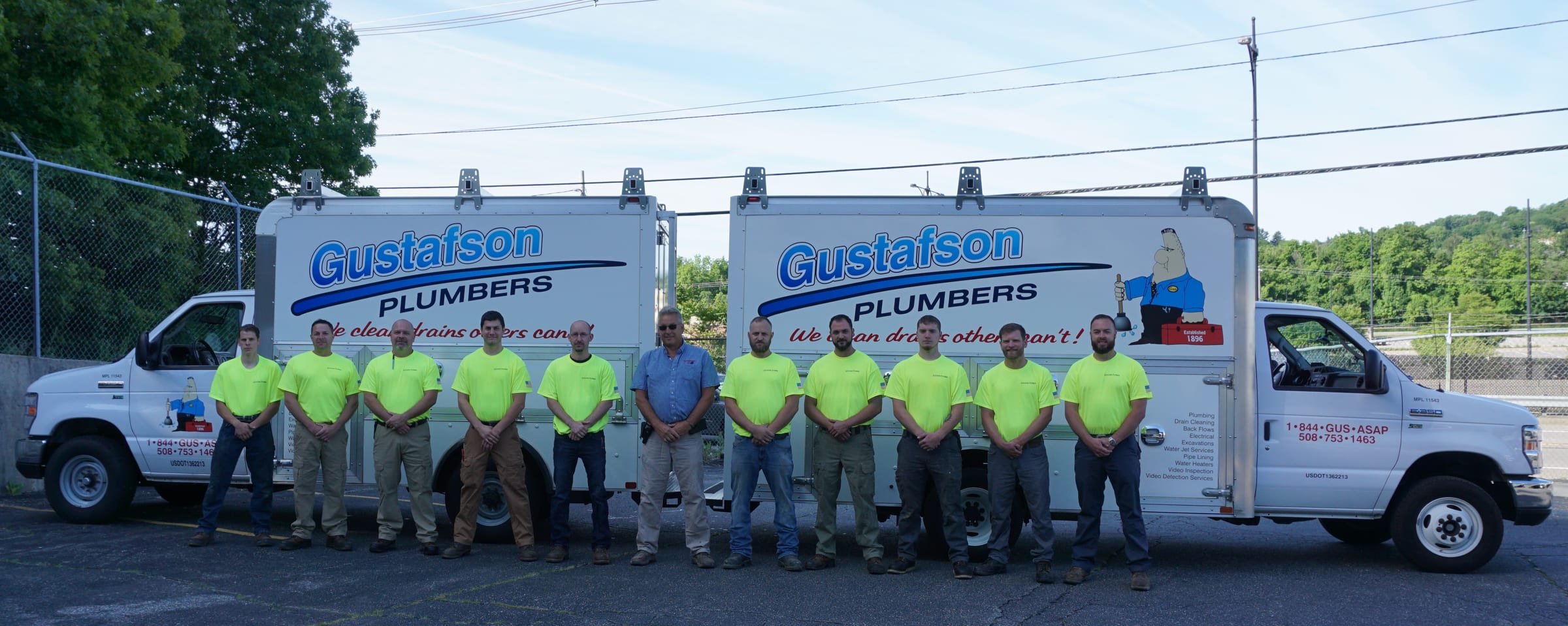 Eleven members of the Gustafson crew, most in hi-vis green shirts, lined up in front of two white Gustafson Plumbers box trucks