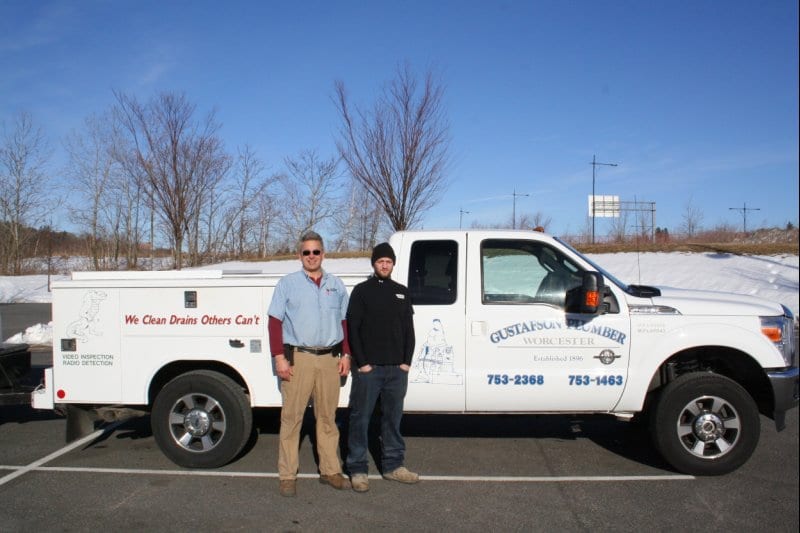 Two men beside an older Gustafson Plumber service pickup lettered Worcester, Established 1896, in a snowy lot
