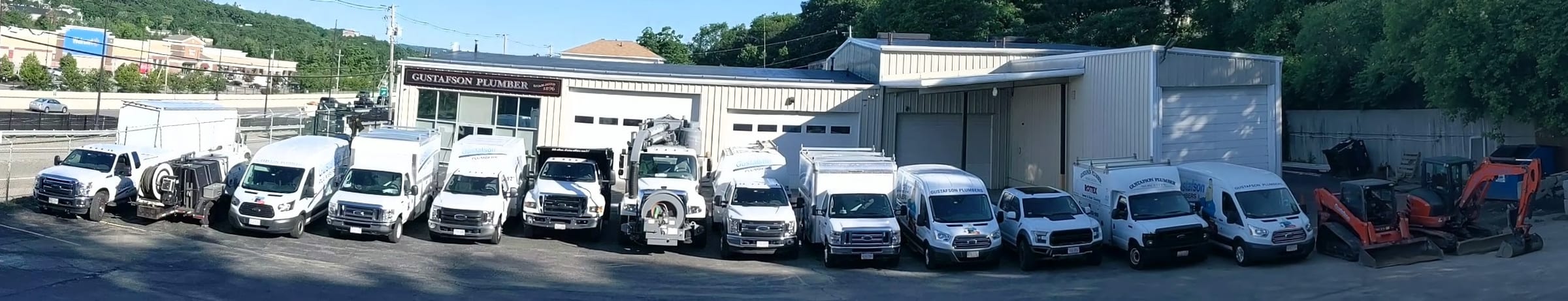 Panorama of the Gustafson fleet of vans, box trucks, a jetter, a vacuum truck and excavators parked in a row in front of the Gustafson Plumber shop