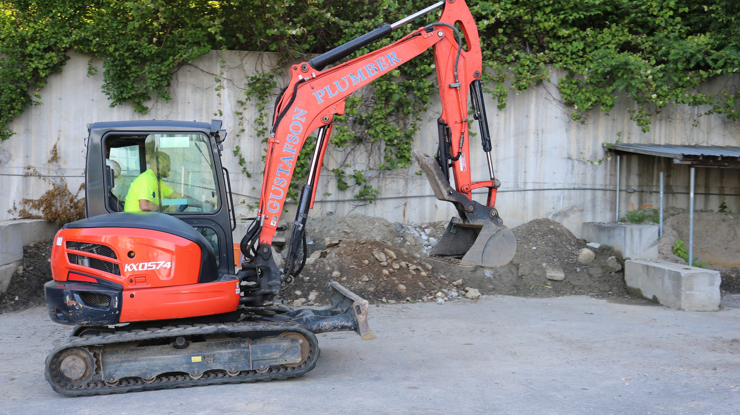 Orange mini excavator lettered Gustafson Plumber, with an operator in hi-vis, digging beside an ivy-covered wall