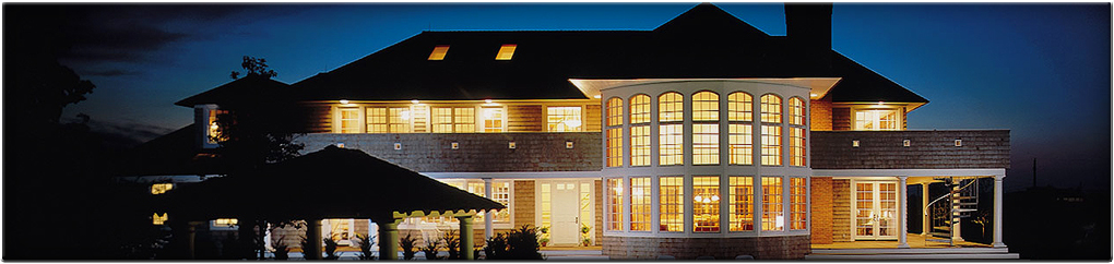 A large shingle-style home glowing with warm interior light at dusk