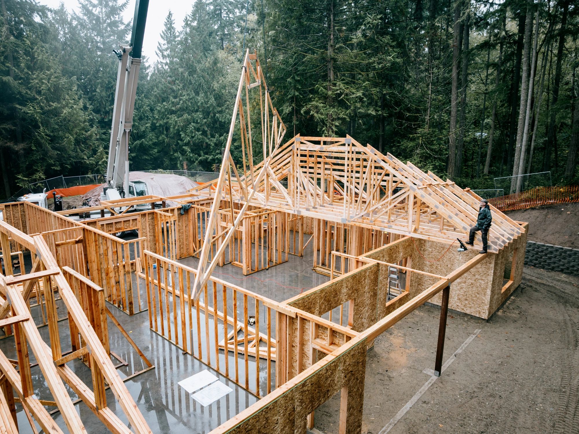 A crane setting roof trusses onto a newly framed single-story house, with stud walls and sheathing in place