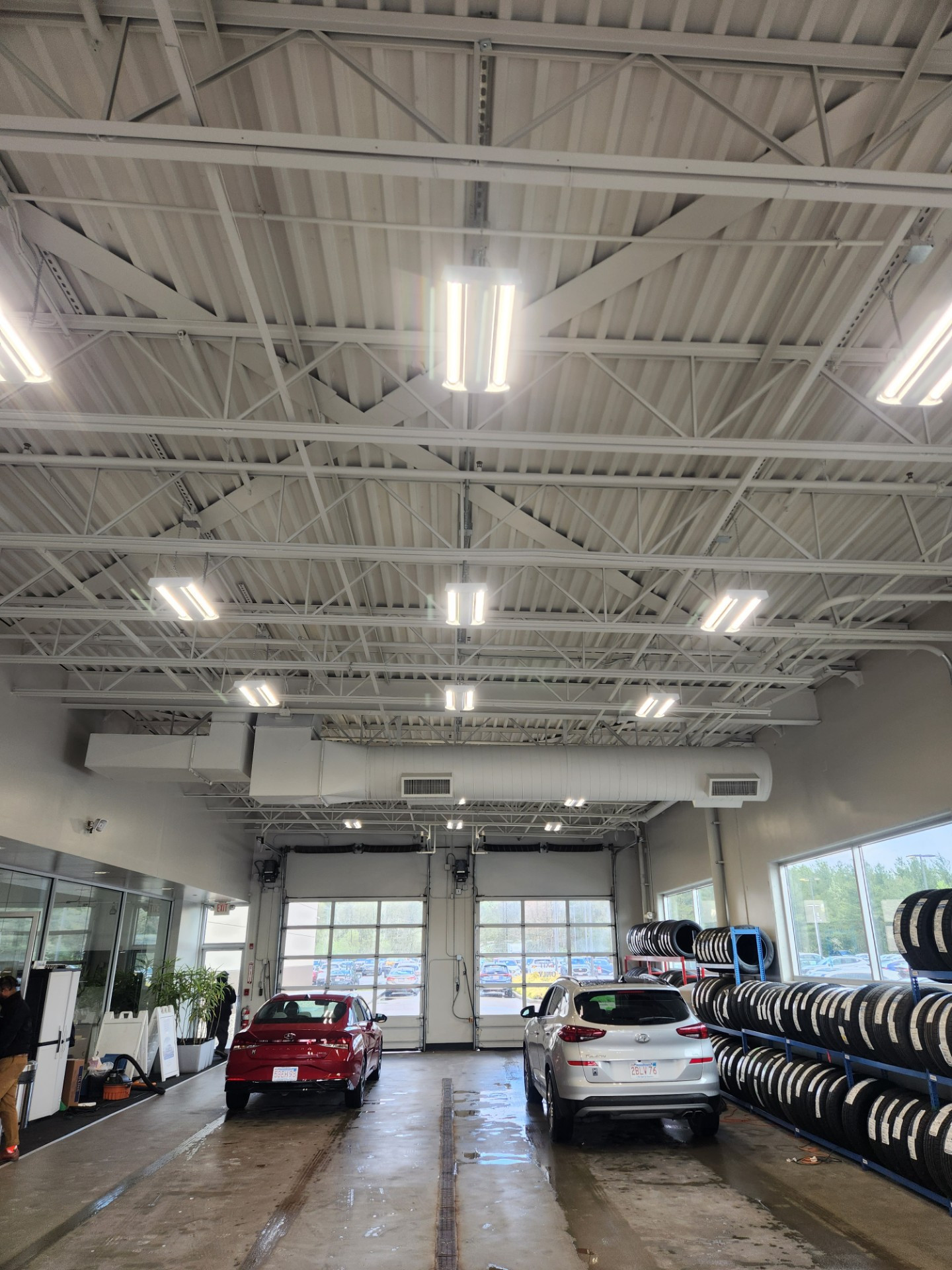 A dealership service drive with linear light fixtures hung in white open-web steel joists above two vehicles and a rack of tires.