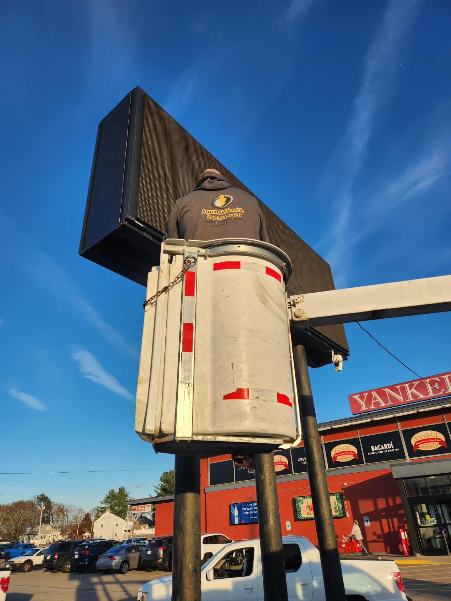 An Ingemi Electric electrician working from a bucket lift at the top of a lighted message board sign against a blue sky.