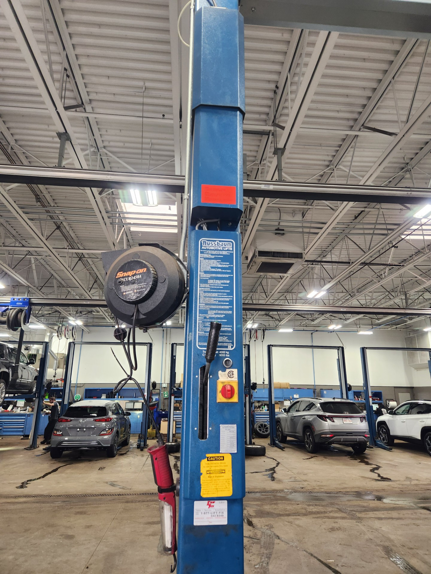 A repair shop floor with blue two-post vehicle lifts and cars, lit by high-bay fixtures in an exposed steel ceiling.