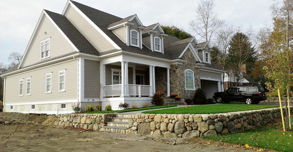 A clapboard-sided home with gabled dormers, a farmer's porch, a stone faced garage bay and a fieldstone retaining wall