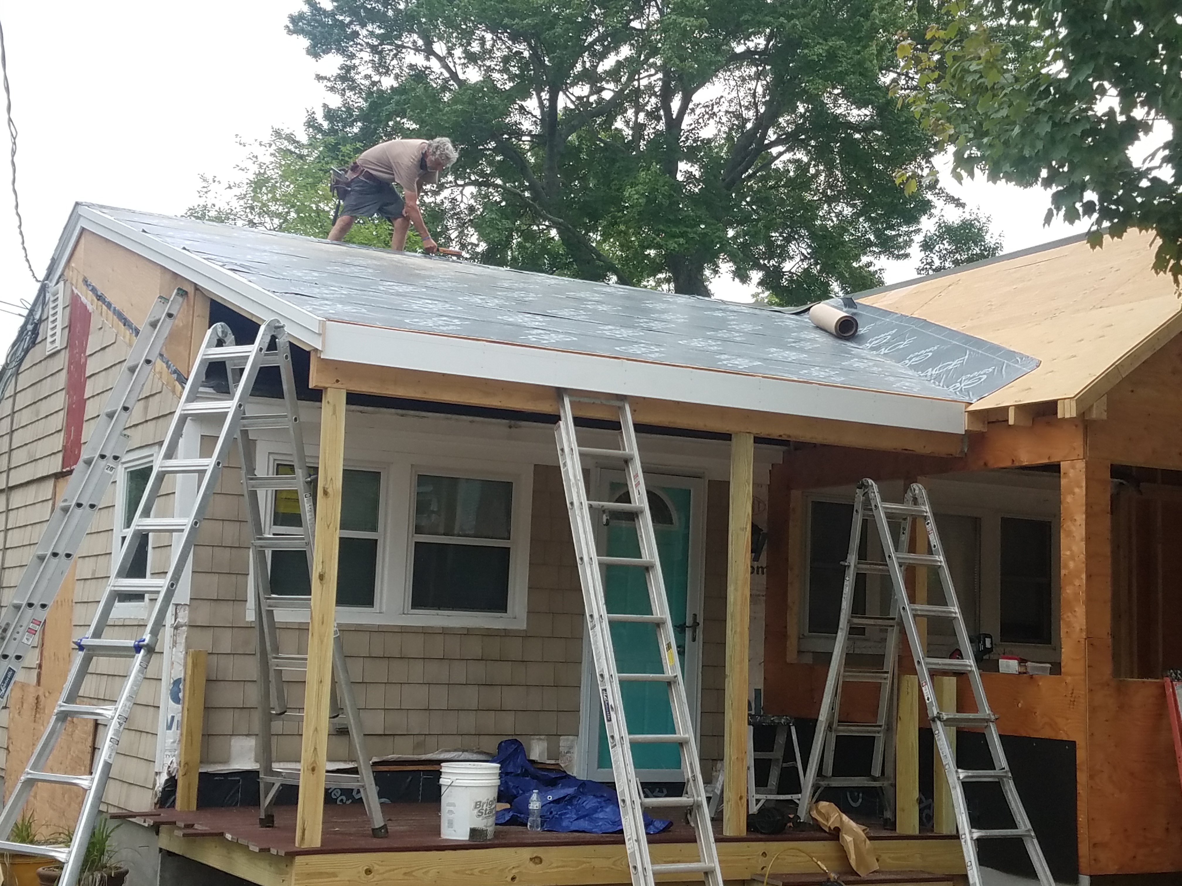 A carpenter on the new porch roof laying underlayment, ladders against the house