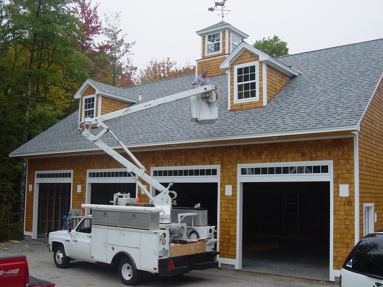 The bucket truck at a large new timber carriage barn, boom raised, a man working from the bucket at the cupola.