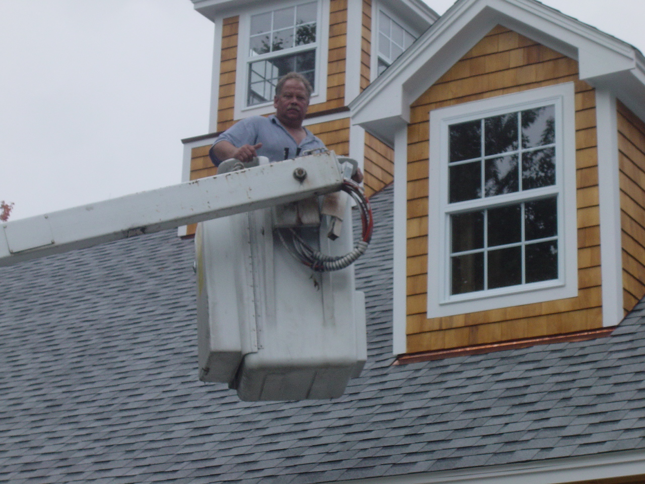 An electrician in the bucket at roof level beside a cedar-shingled dormer, a coil of cable on the bucket rail.