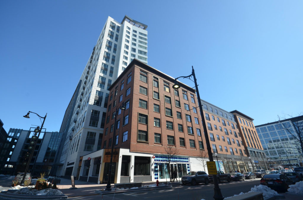 A finished high-rise tower rising behind a brick mid-rise on a city block under a clear winter sky.