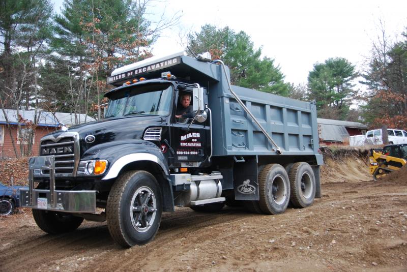 A black Mack tri-axle dump truck lettered Miles Of Excavating, driver in the cab, on a dirt site