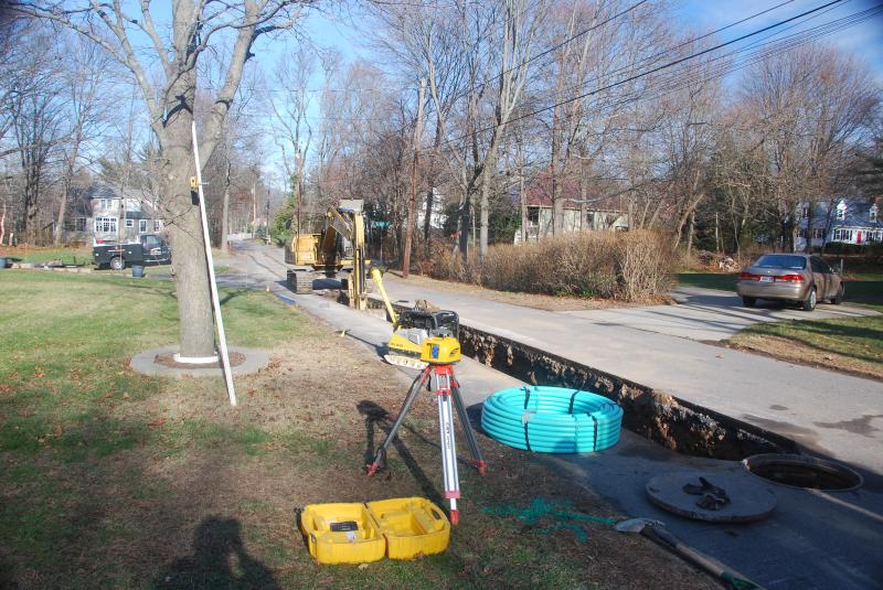 An open water service trench along a residential street, with coiled blue poly pipe, a laser level on a tripod and a machine working at the end of the road