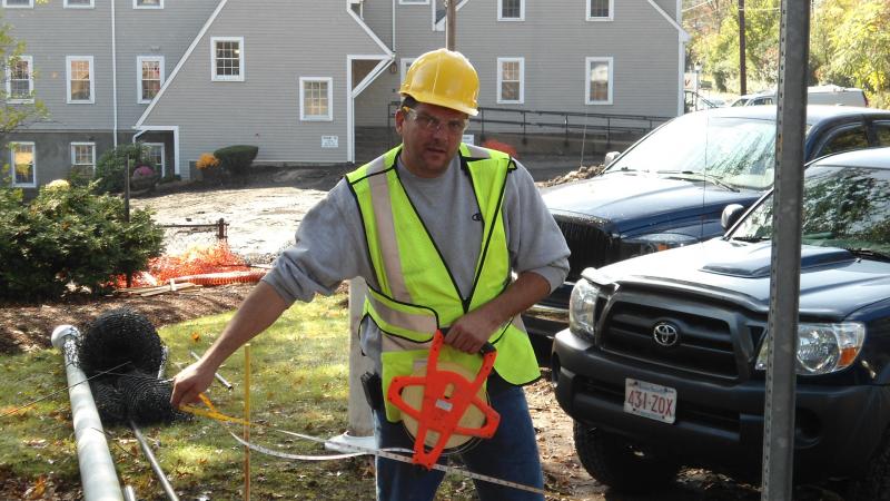 A worker in a hard hat and hi-vis vest running a pipe locator beside a pickup truck