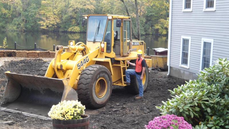 A John Deere 544G wheel loader working close beside a house, its siding and flowerbeds a few feet from the bucket