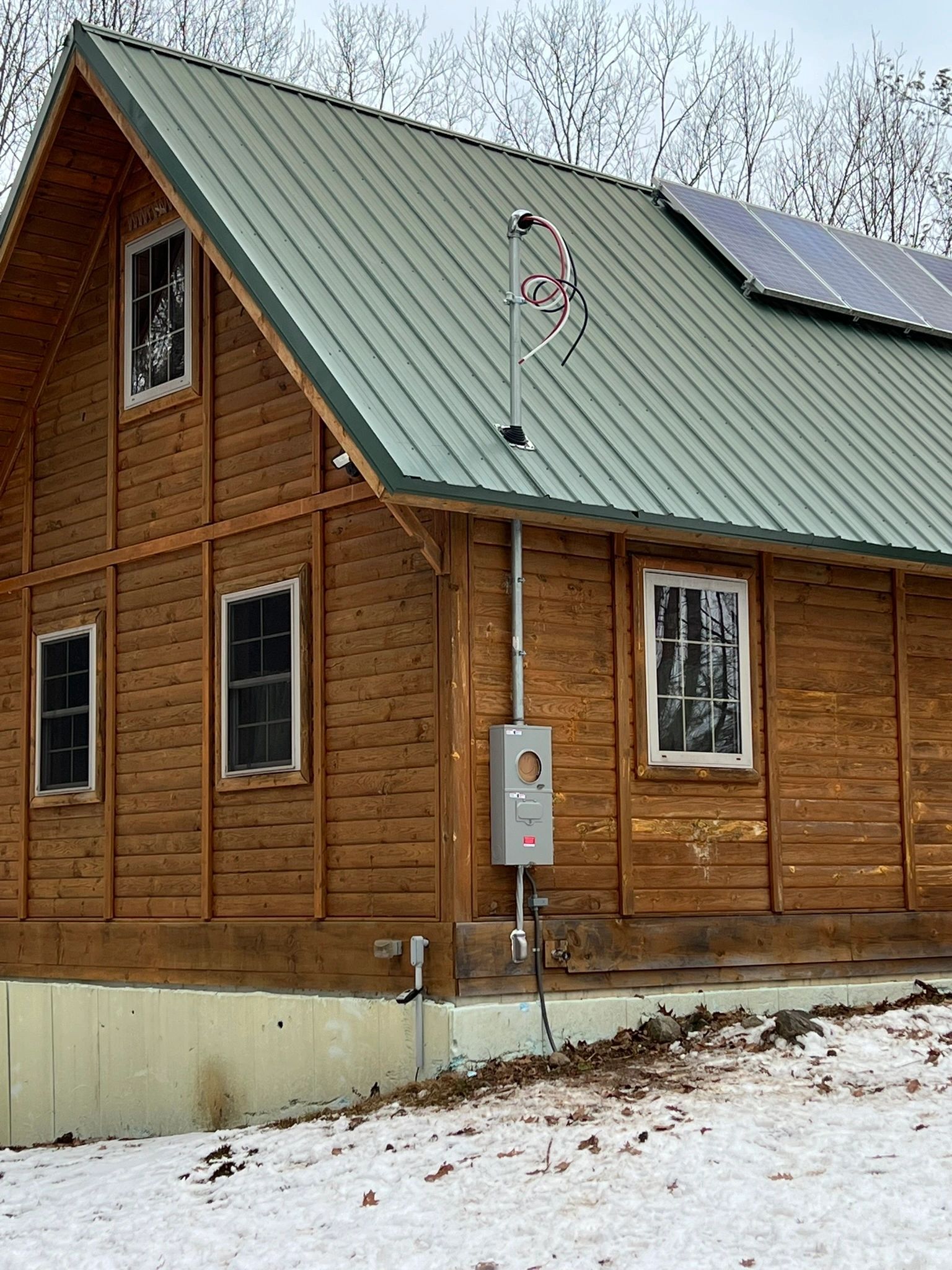 A log home with a green standing seam roof, with a service mast, meter and disconnect on the gable wall, in snow