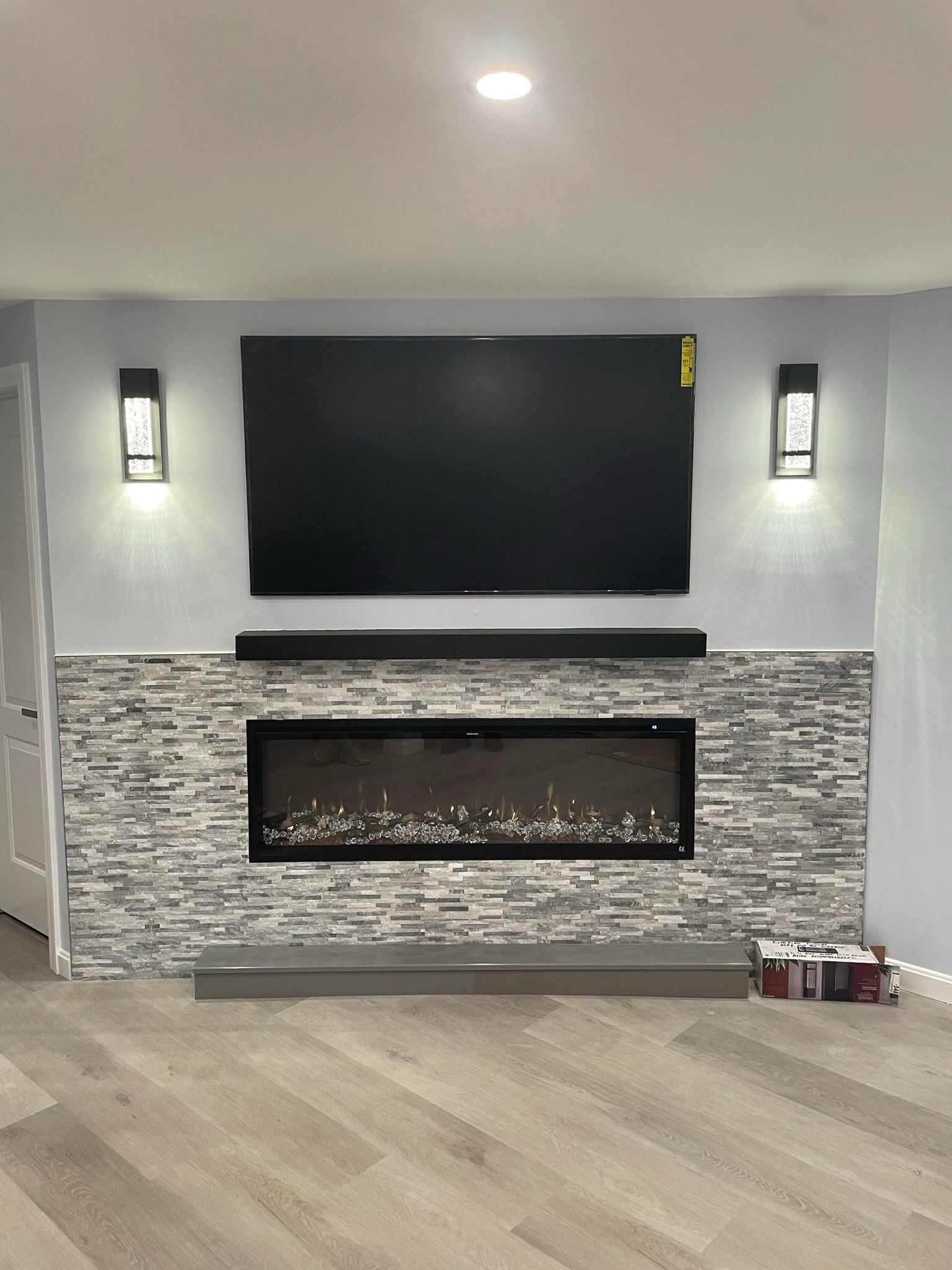 A finished living room wall with a wall mounted television above a linear fireplace in stacked stone, flanked by two lit sconces