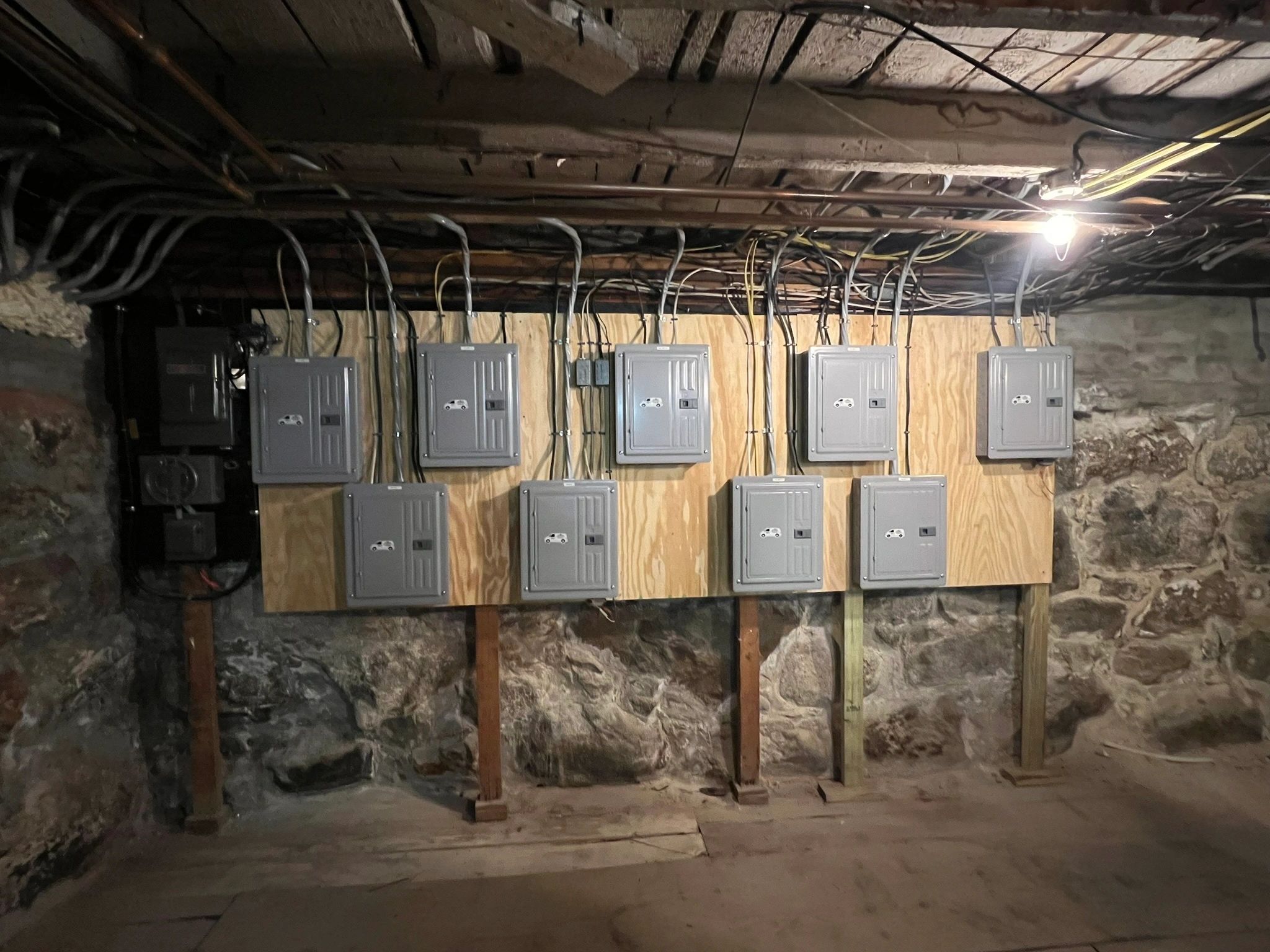 A bank of electrical panels and disconnects mounted on plywood against an old fieldstone foundation wall in a dim basement