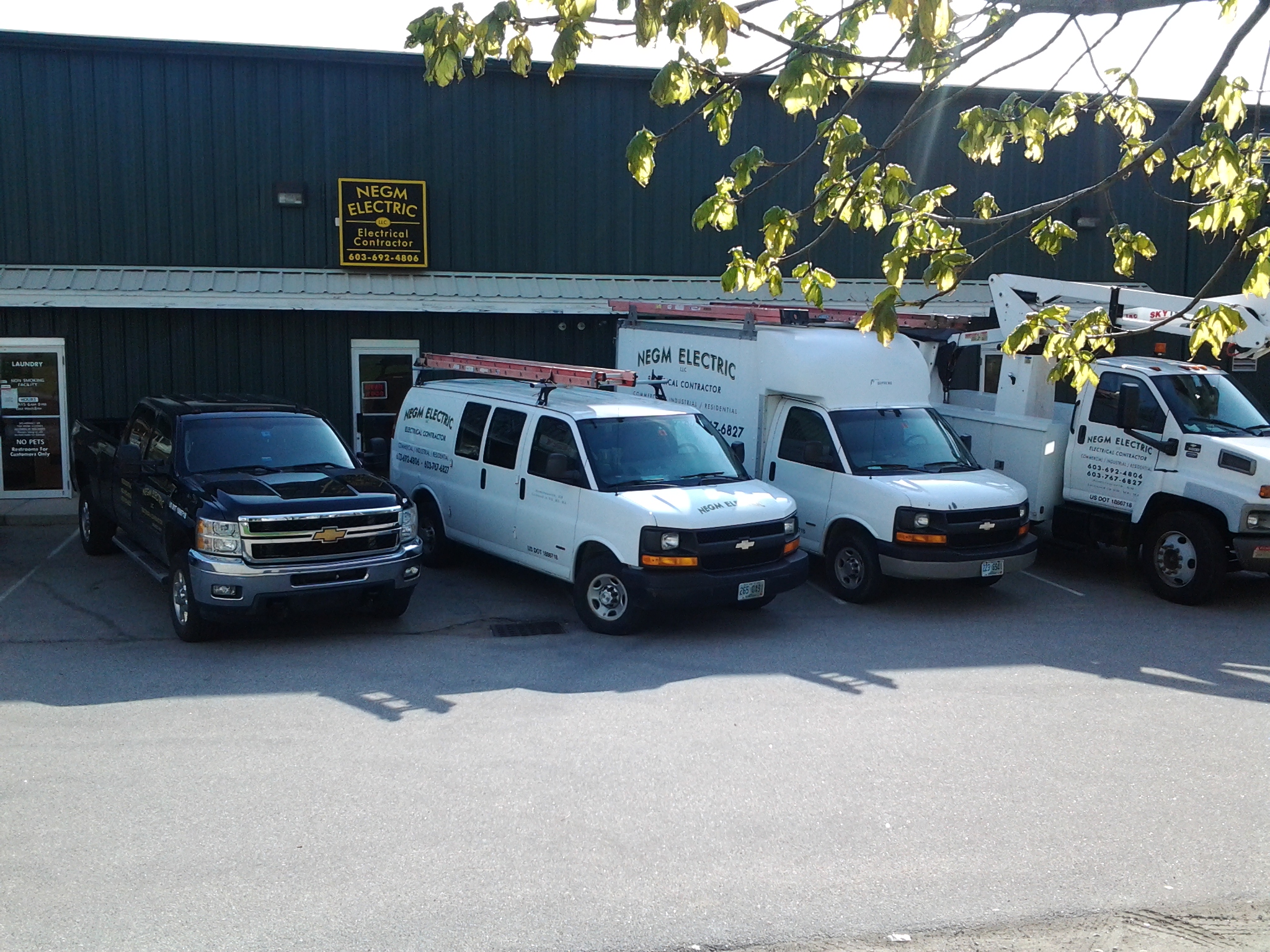 The Negm Electric yard on a bright day: three white service vans, a bucket truck and a black pickup parked along a dark green metal building with the NEGM Electric sign over the door.