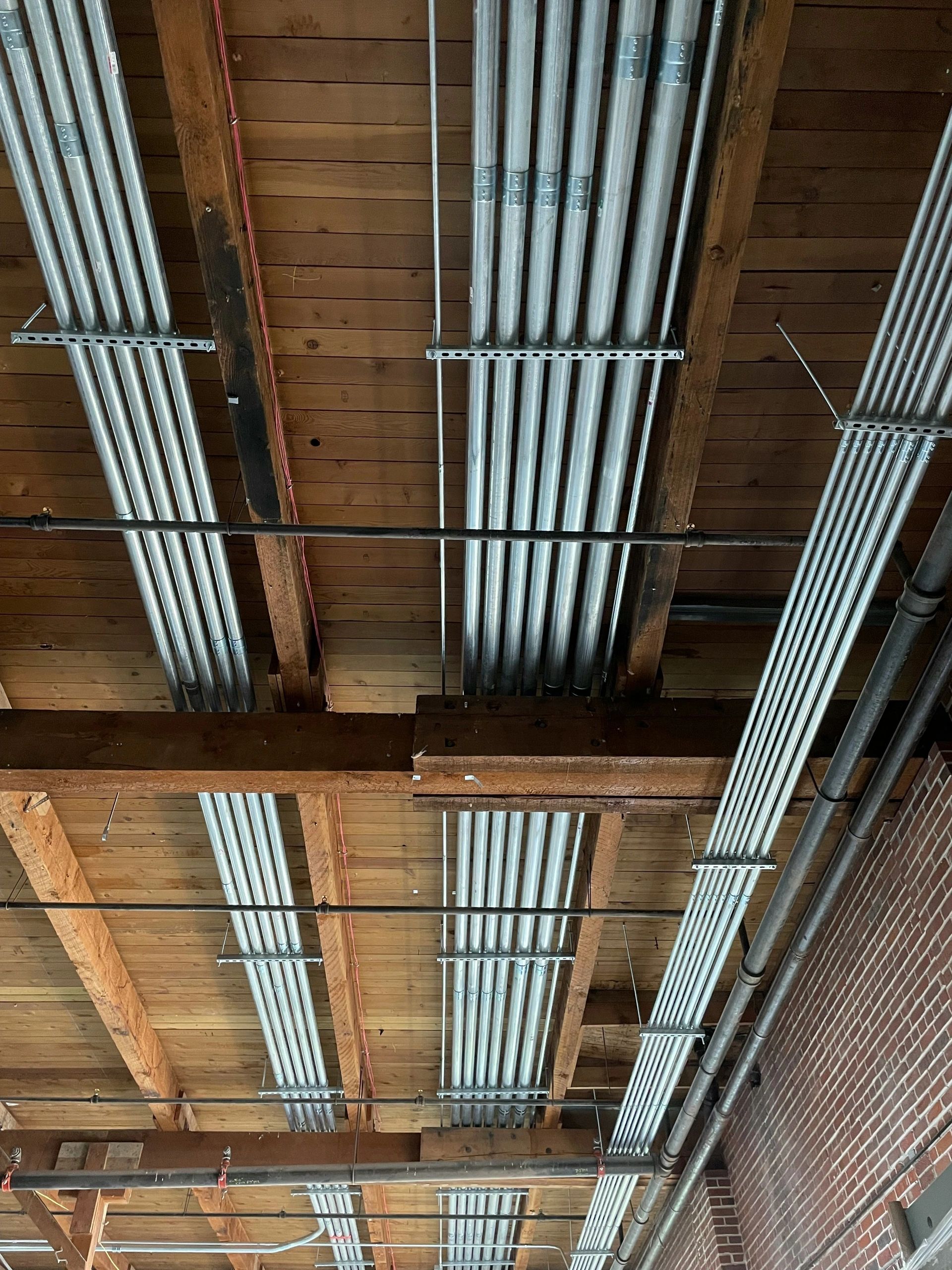 Parallel runs of galvanized conduit and fluorescent strip fixtures mounted to the wood-beam ceiling of a mill building.