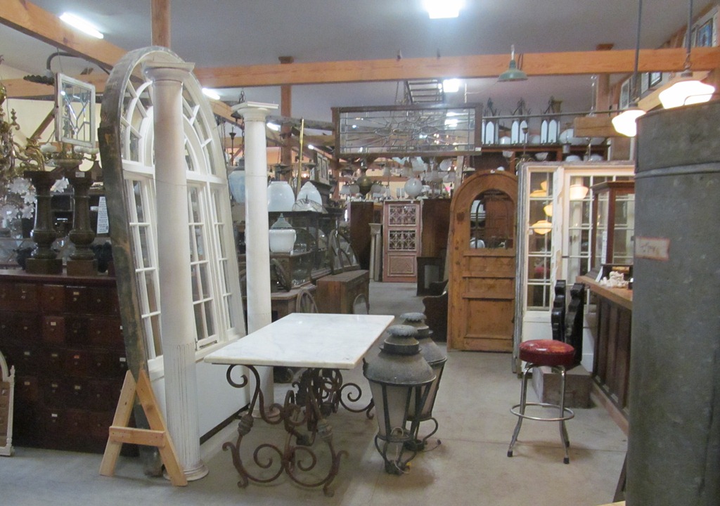 Showroom floor at Nor'East Architectural Antiques with an arched sash window, a pair of salvaged columns, a round table on an ornamental base and a tall panelled door.
