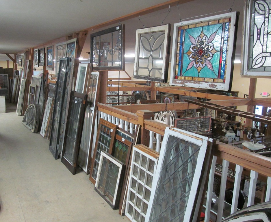 Rows of salvaged sash windows stacked along a warehouse wall, with leaded and stained glass panels hung above them.