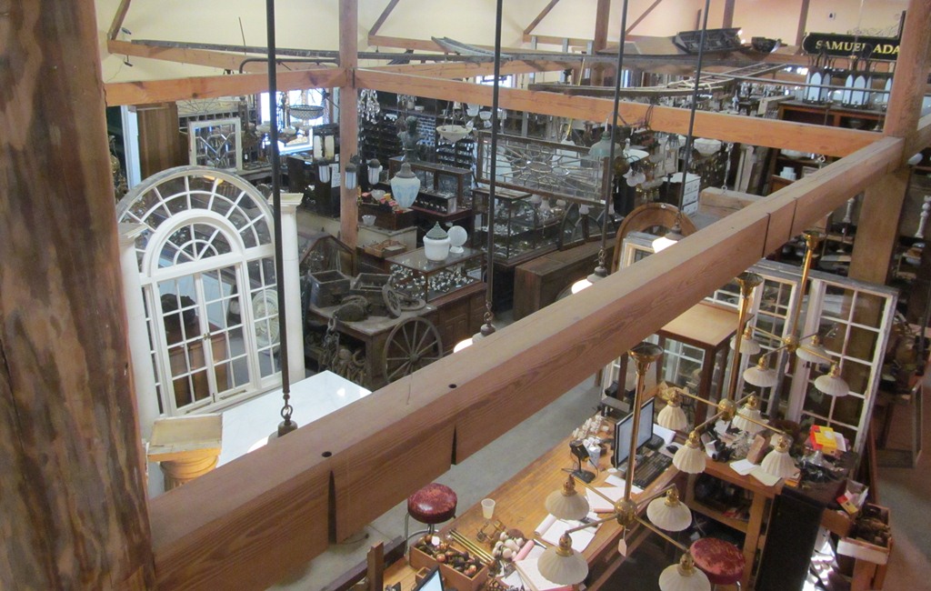 View from the timber-framed loft rail down onto the showroom floor, showing an arched window, cases of small antiques and hanging lamps.