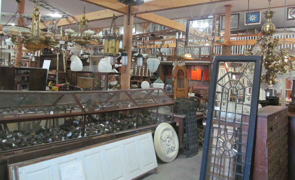 Chandeliers and glass shades hanging above a long display case filled with salvaged door hardware.