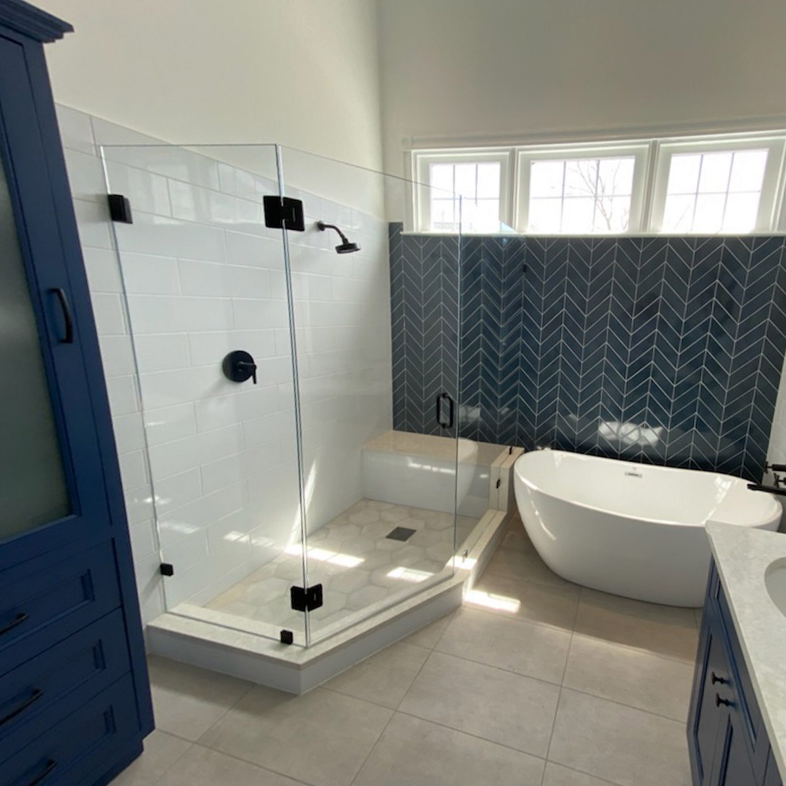 The same bathroom after work, with a glass shower, blue chevron tile, a freestanding tub and a navy vanity