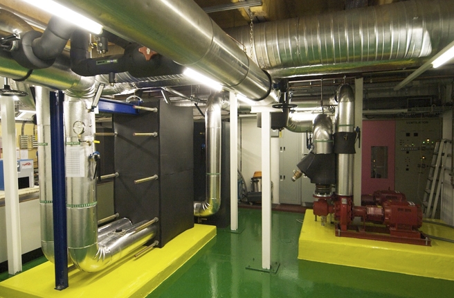 A mechanical room with a plate heat exchanger, insulated duct and red pumps on a painted plant floor