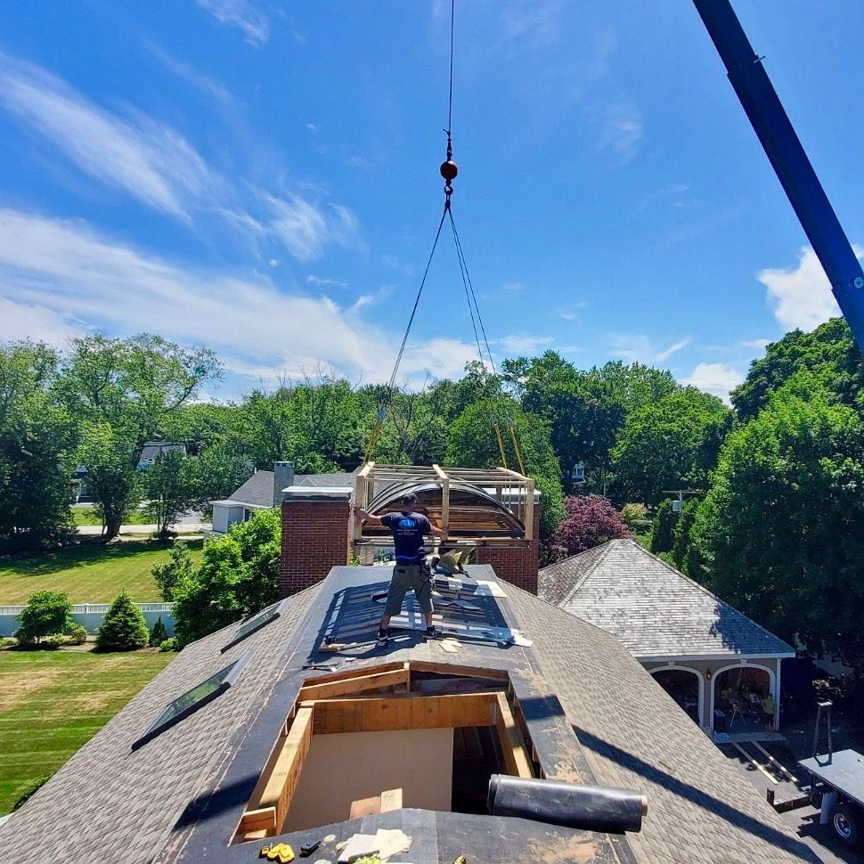 A crane boom over a roof mid lift with a green field beyond