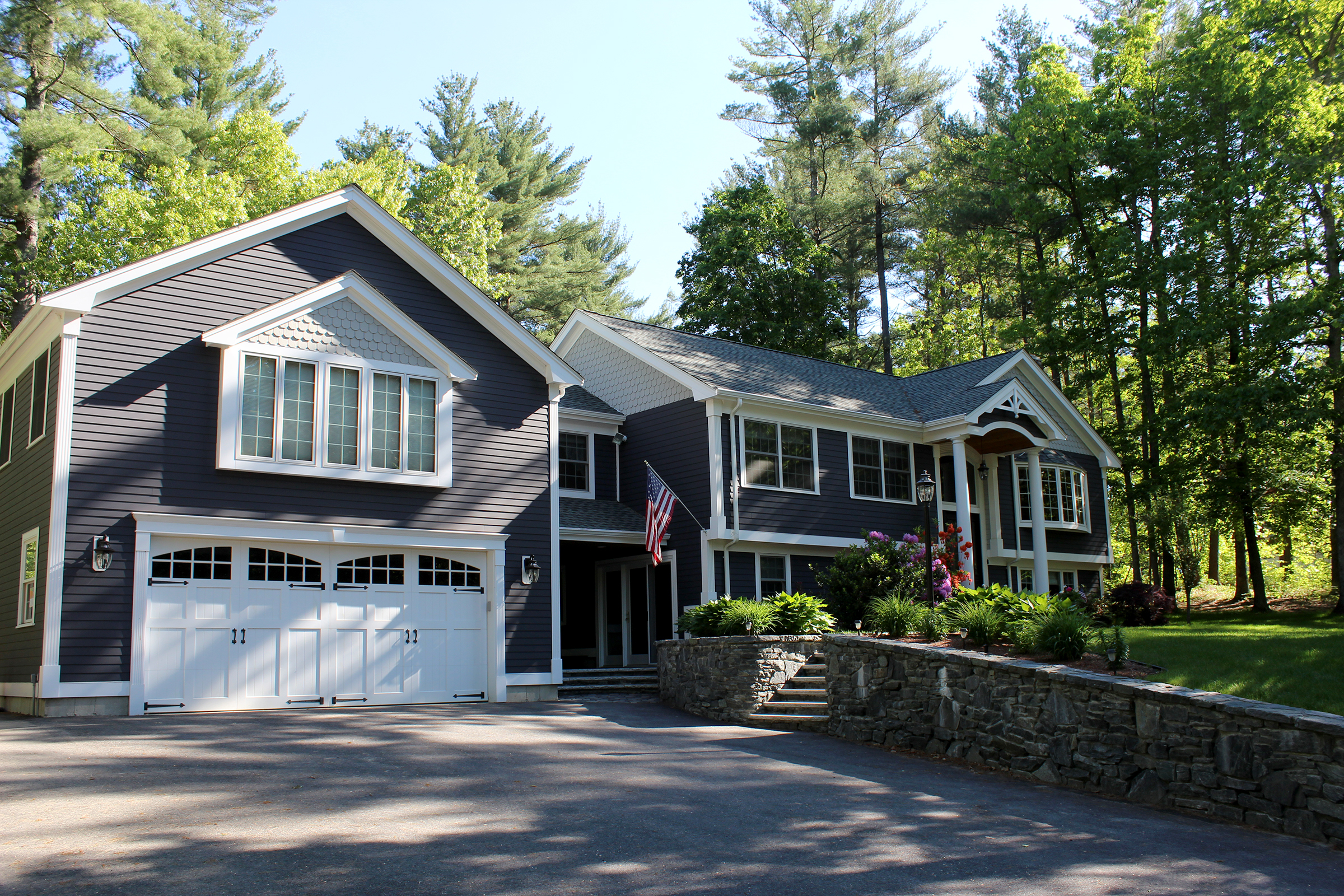 Remodeled raised ranch in dark blue siding with a two-car garage wing, white trim, a columned entry and a fieldstone wall