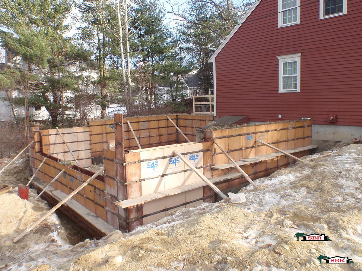Foundation forms set for an addition beside a red colonial in winter