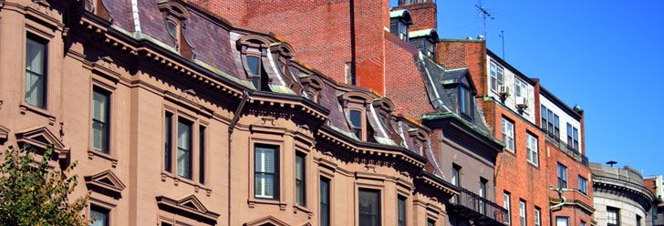 Brick and brownstone rowhouse facades with mansard roofs and dormer windows under a clear sky in greater Boston.