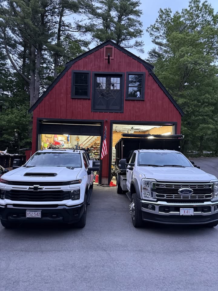 A red gambrel-roof barn at dusk with both bay doors open and an American flag between them, two white work trucks parked in front.